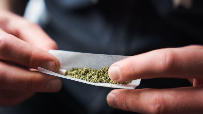 Close-up of a young adult rolling a marijuana joint against a blurred background. Man placing desiccated marijuana leaves inside rolling paper.