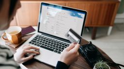 Over the shoulder view of young Asian woman shopping online for flight tickets on airline website with laptop, entering credit card details to make mobile payment at home. Camera and passport on the table. Travel planning. Booking a holiday online