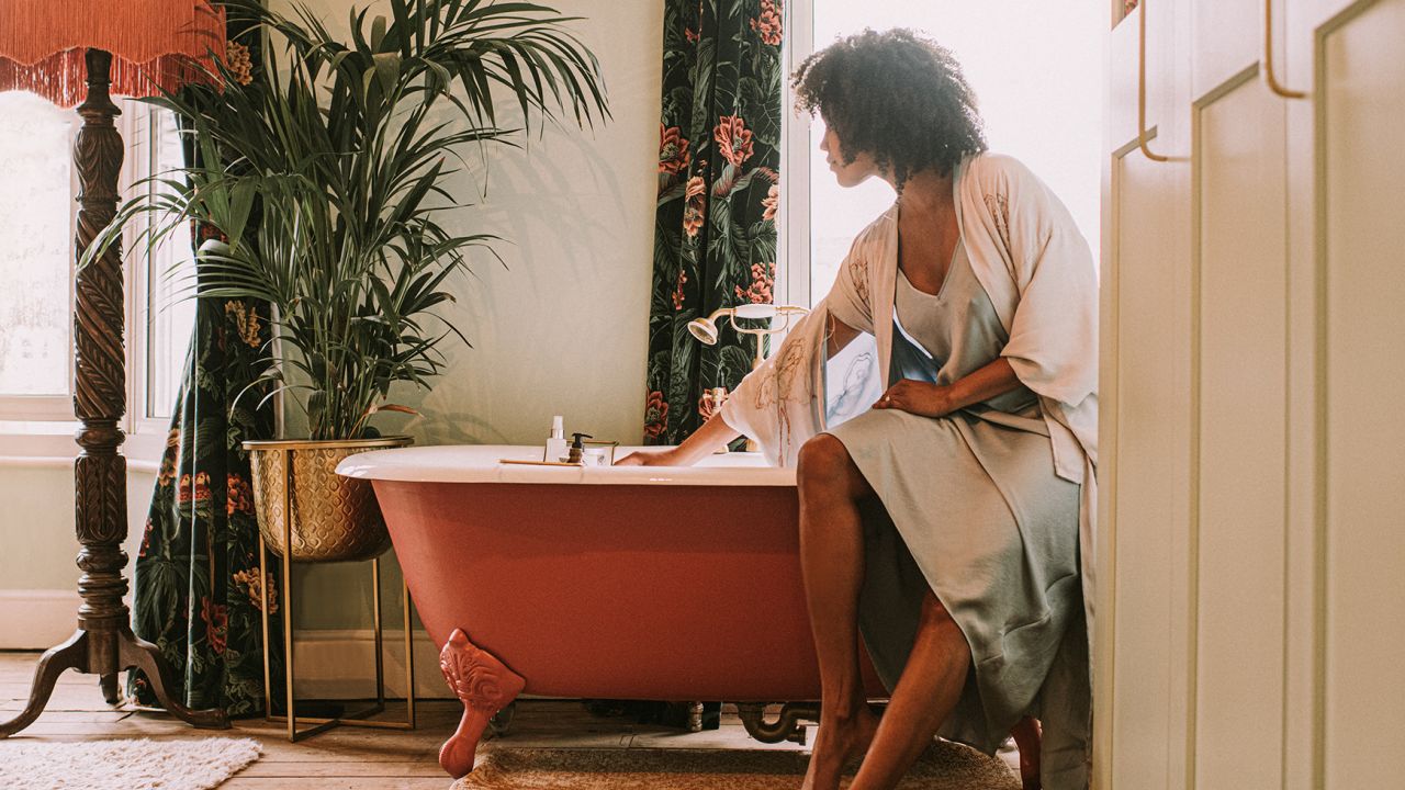 A beautiful woman perches on the side of an elegant red roll top bathtub, and waits for it to fill up. She wears a silky robe. Light floods through the window, backlighting the relaxing scene and giving it a dreamy vibe.