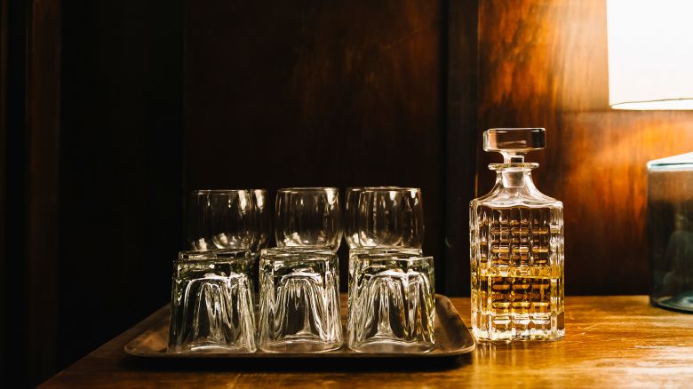 An ornate glass bottle of half empty whisky  with a set of glasses and tumblers on a serving tray, in atmospherically lit on an old-fashioned, classy wood-panelled bar.