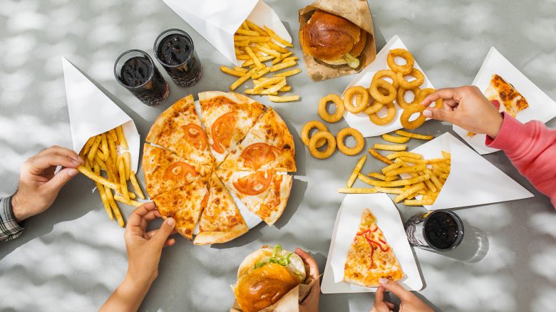 Flat-lay of friends group eating burgers, fries, onion rings, pizza, drinking cola at outdoor party over table background, top view. Fast food dinner from delivery service.Fast food and unhealthy eating concept