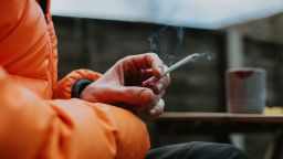 A man sits outside and holds a slim menthol cigarette between his fingers. Close-up with focus on the cigarette.