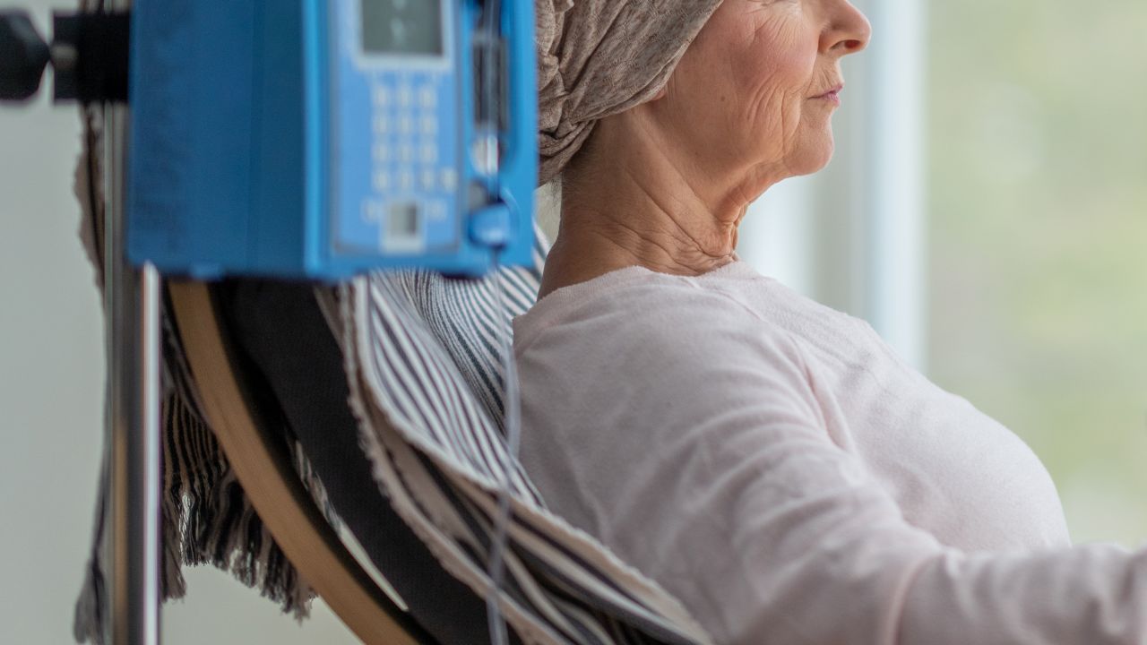 A senior woman sits in the comfort of her own home as she receives her chemotherapy treatment.  She is dressed comfortably and wearing a headscarf to keep her warm.