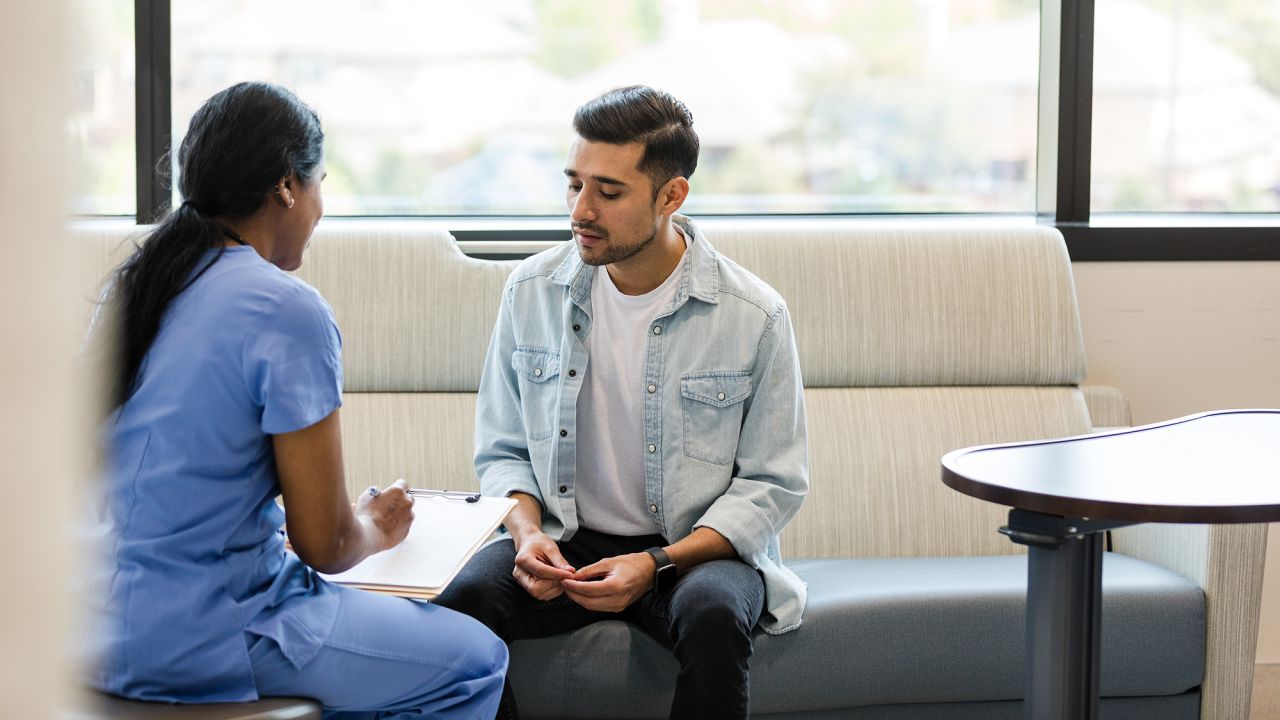 The young adult male sits on the hospital room couch to talk with the hospital insurance specialist to update medical information.