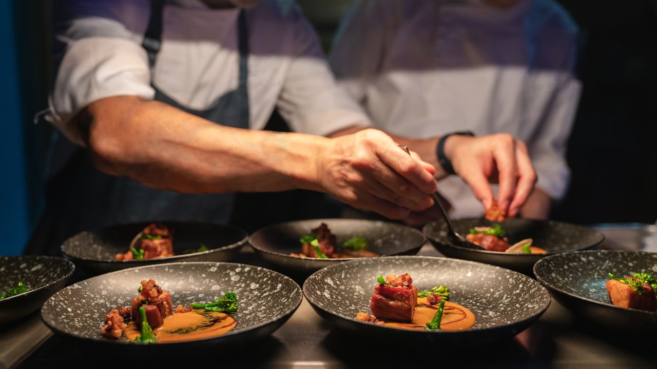 Chefs preparing food in professional kitchen. Focus on dish of dinning food in front row. Chef blur as background.
