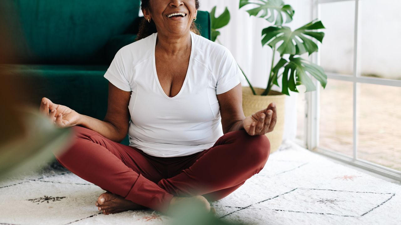 Black elderly woman meditating in lotus position at home, wearing a happy smile. Retired senior prioritizing her mental health and wellbeing, finding peace and serenity in her yoga practice.