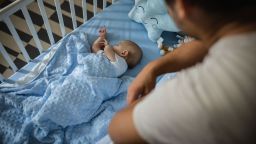 Cute silent little baby boy in white babysuite is in his crib, lying on his back, covered with blue blanket. He is sleeping while his father taking care of him. Love emotion.