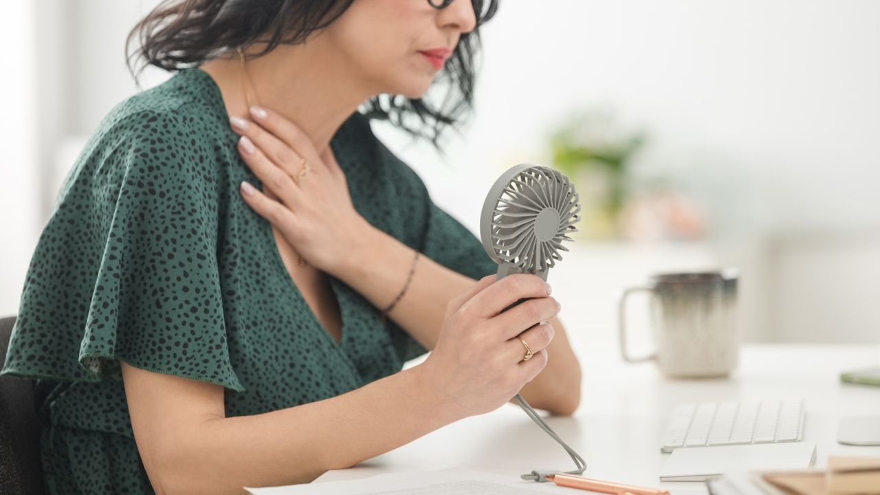 Menopausal mature woman wearing green dress sitting at the desk in the office, having hot flashes and using hand fan.