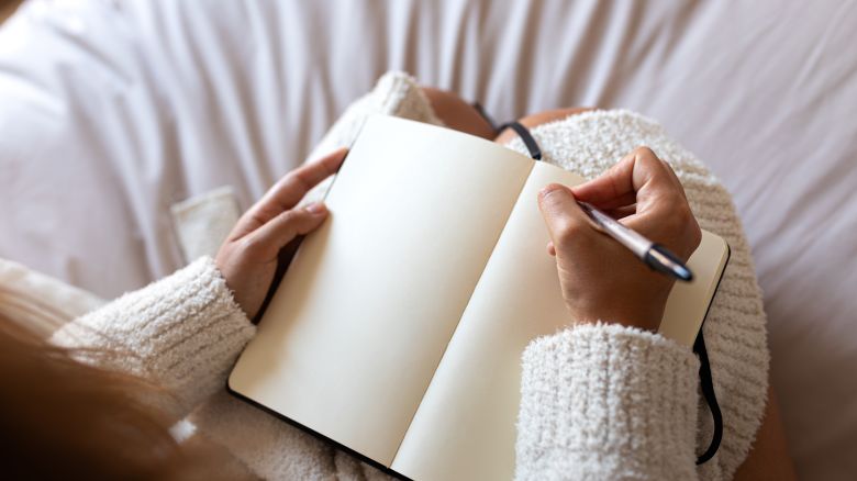 POV high angle view of Unrecognizable young woman wearing a robe sitting on bed writing on journal in cozy bedroom. Lifestyle concept.