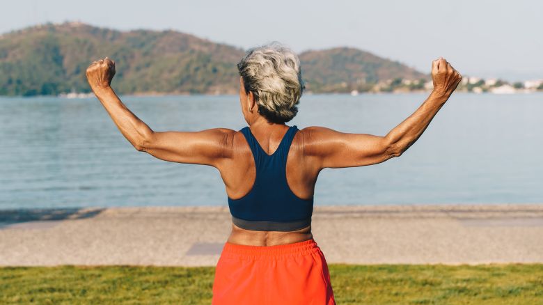 Senior gray-haired woman doing exercises in public park, sunny summer morning. Rear view.