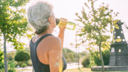 An elderly gray-haired woman in a sports top drinks water after working out in the park. Sunny summer morning.