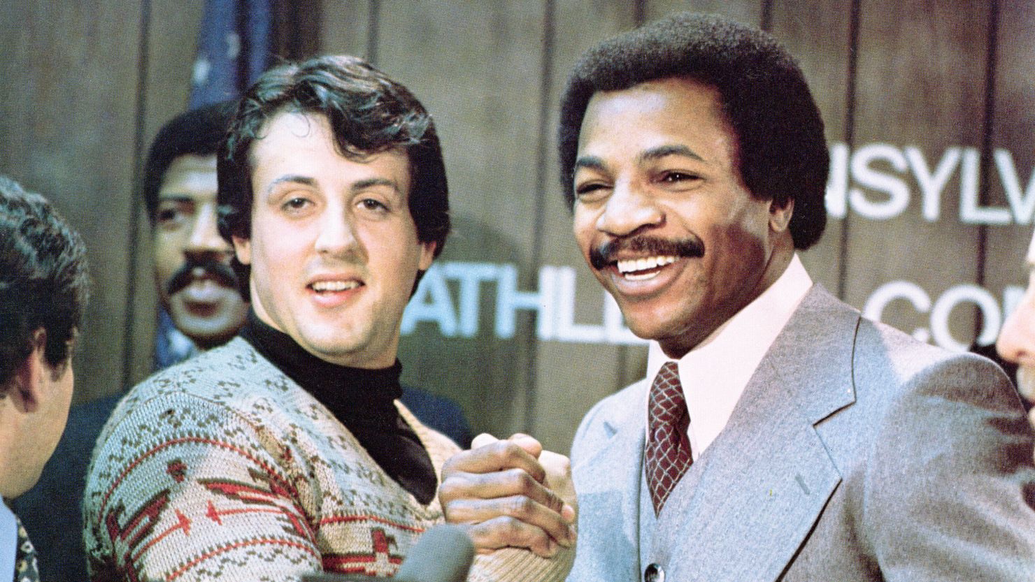 Sylvester Stallone and Carl Weathers grip hands and smile together during a press conference in a still from the film, 'Rocky,' directed by John G. Avildsen, 1976.