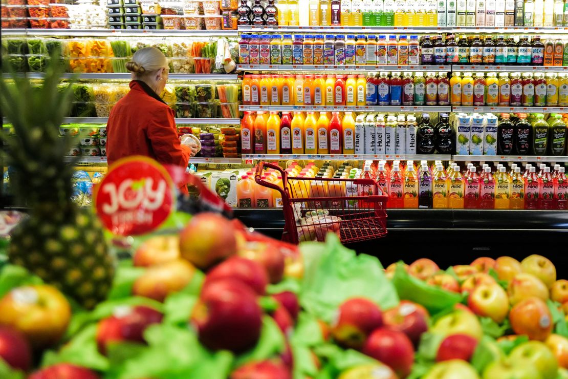 A woman in a red coat stands in front of a grocery store stand of fruits, vegetables and juice. 