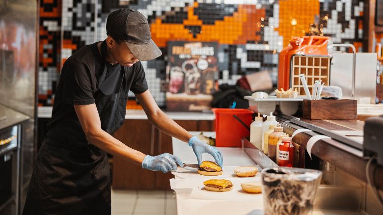 Man, burger and fast food kitchen worker at counter with gloves, cooking and uniform at small business. Restaurant, sandwich and cook with hamburger patty stack for lunch service with take away.