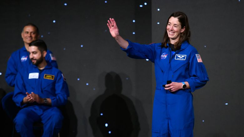 A woman with dark hair, wearing a blue NASA astronaut jumpsuit, waves and smiles. 