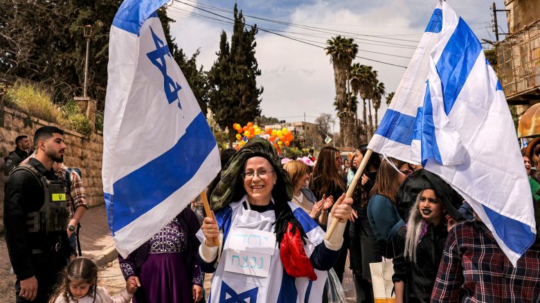 Israeli politician Orib Strook stands amidst a small crowd, holding two Israeli flags. She is wearing a third flag, draped over her body. 