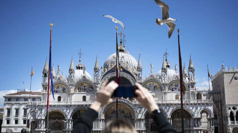 A woman uses her smartphone to take footage of the Basilica with flying gulls in San Marco Square in Venice, on April 25, 2024. The new strategy to lower the number of tourists visiting the UNESCO World Heritage site calls for day-trippers to pay a five-euro ticket to enter the historic city centre and is due to start on April 25. (Photo by MARCO BERTORELLO / AFP) (Photo by MARCO BERTORELLO/AFP via Getty Images)