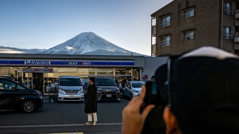 This photo taken on January 1, 2024 shows a tourist posing in front of a convenience store with Mount Fuji in the background, in the town of Fujikawaguchiko, Yamanashi prefecture. A huge black barrier to block Mount Fuji from view will be installed in a popular photo spot by Japanese authorities exasperated by crowds of badly behaved foreign tourists, it was reported on April 26, 2024. (Photo by Philip FONG / AFP) (Photo by PHILIP FONG/AFP via Getty Images)
