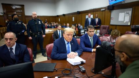 Donald Trump sits at a table amidst a crowded court room.
