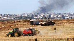 An agricultural vehicle harvests hay in front of an Israeli army battle tank in southern Israel near the border with the Gaza Strip, as smoke rises above the Palestinian territory on May 15, 2024, amid the ongoing conflict between Israel and the Hamas movement.