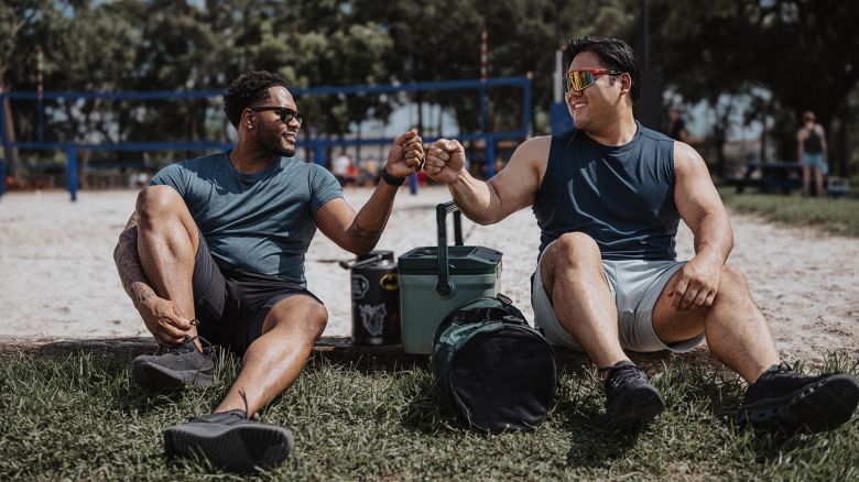 A black man and Asian man sit in the grass next to a beach volleyball court located at a park in Houston, Texas, and greet each other with a fist bump.