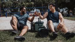 A black man and Asian man sit in the grass next to a beach volleyball court located at a park in Houston, Texas, and greet each other with a fist bump.
