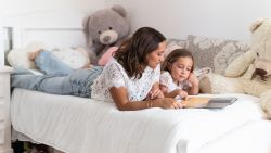 Girl and mother reading picture book lying on bed next to two teddy bears