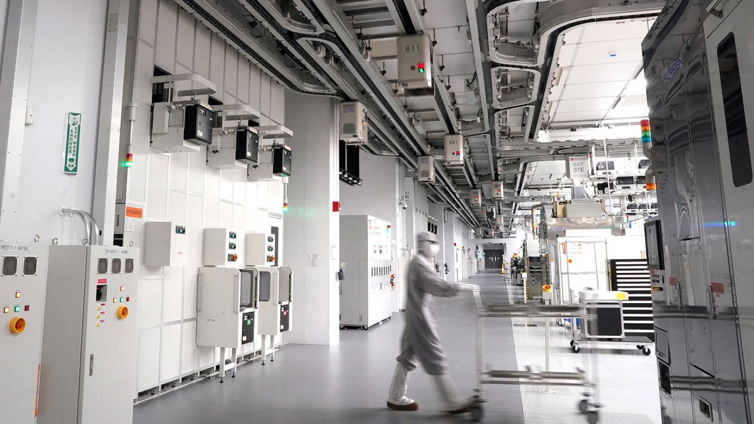 An employee wearing a cleanroom suit walks beneath Automated Material Handling Systems (AMHS) vehicle robots moving along tracks on the ceiling inside the GlobalFoundries semiconductor manufacturing facility in Malta, New York, US, on Tuesday, June 18, 2024.