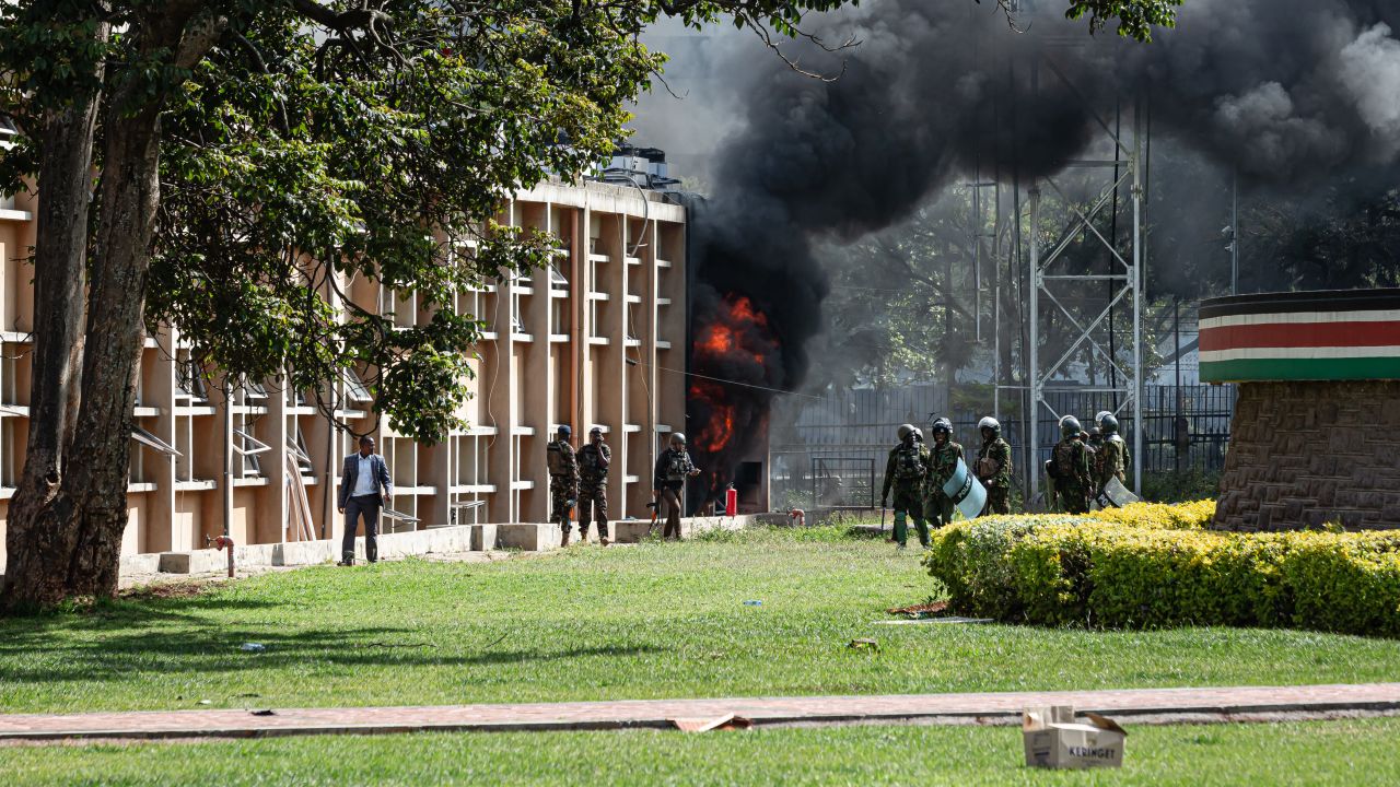 NAIROBI, KENYA - JUNE 25: Kenyan police officers stand as fire comes out of the parliament building during a protest against the finance bill on June 25, 2024 in Nairobi, Kenya. Last week saw several days of protests, mainly by young Kenyans, against a proposed finance bill that promises to raise taxes on a variety of goods. The outcry spurred the government to withdraw several contentious provisions, including taxes on bread and vehicles, but the bill passed a second-round vote and a parliamentary committee is now considering amendments. (Photo by Patrick Meinhardt/Getty Images)