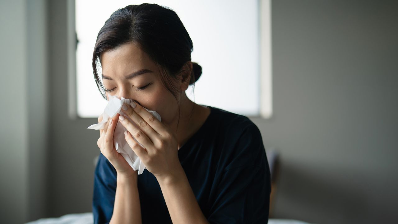 Young Asian woman feeling ill, suffering from cold, flu and allergy, blowing her nose covered with a facial tissue. Concept of health and illness