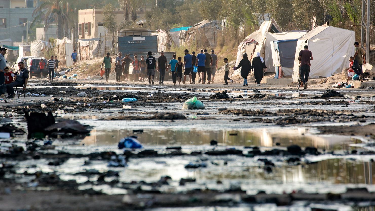 Palestinians walk near tents used as shelter along a street covered with stagnant wastewater in Deir el-Balah, in the central Gaza Strip on July 19, 2024.