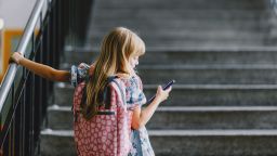 A young girl with a backpack holds her phone while standing on a school staircase. Back to school concept.