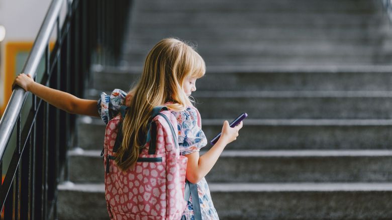 A young girl with a backpack holds her phone while standing on a school staircase. Back to school concept.