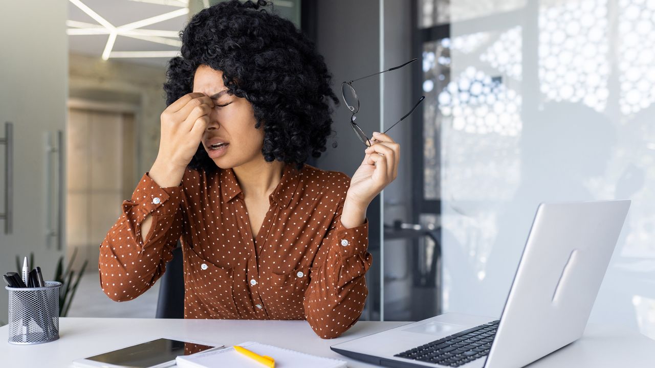 Stressed businesswoman holding head, feeling headache while working at office desk with laptop and documents. Business stress and fatigue concept.