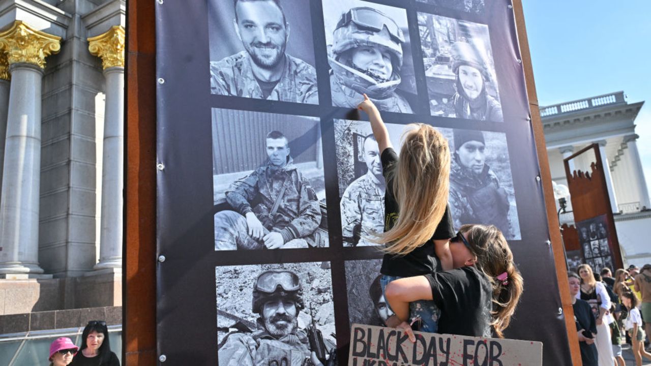 A mother holds her daughter who points the portrait of her late father at an exhibition in the sidelines of a memorial event at Independence Square in Kyiv on July 28, 2024, marking second year anniversary of the Olenivka prison attack. On July 29, 2022, dozens of Ukrainian soldiers died in the bombardment of the Russian-controlled jail of Olenivka in the eastern Donetsk region. (Photo by Sergei SUPINSKY / AFP) (Photo by SERGEI SUPINSKY/AFP via Getty Images)