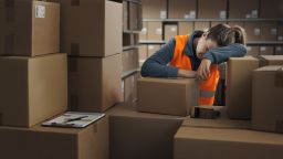 Exhausted warehouse worker leaning on a pile of boxes and sleeping at work