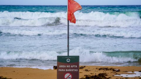 A warning sign reading "Danger, No Swimming" under a red flag, indicating unsafe swimming conditions, are seen on the beach as waves crash onto the shore in Luquillo, Puerto Rico, on August 13, 2024, as Tropical Storm Ernesto approaches. Ernesto is about 300 miles (480kms) east southeast of Puerto Rico, according to the US National Hurricane Center and predicts the storm will reach the island by late August 13. (Photo by Jaydee Lee SERRANO / AFP) (Photo by JAYDEE LEE SERRANO/AFP via Getty Images)
