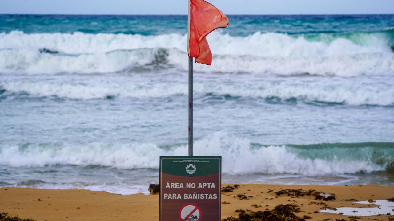 A warning sign reading "Danger, No Swimming" under a red flag, indicating unsafe swimming conditions, are seen on the beach as waves crash onto the shore in Luquillo, Puerto Rico, on August 13, 2024, as Tropical Storm Ernesto approaches. Ernesto is about 300 miles (480kms) east southeast of Puerto Rico, according to the US National Hurricane Center and predicts the storm will reach the island by late August 13. (Photo by Jaydee Lee SERRANO / AFP) (Photo by JAYDEE LEE SERRANO/AFP via Getty Images)