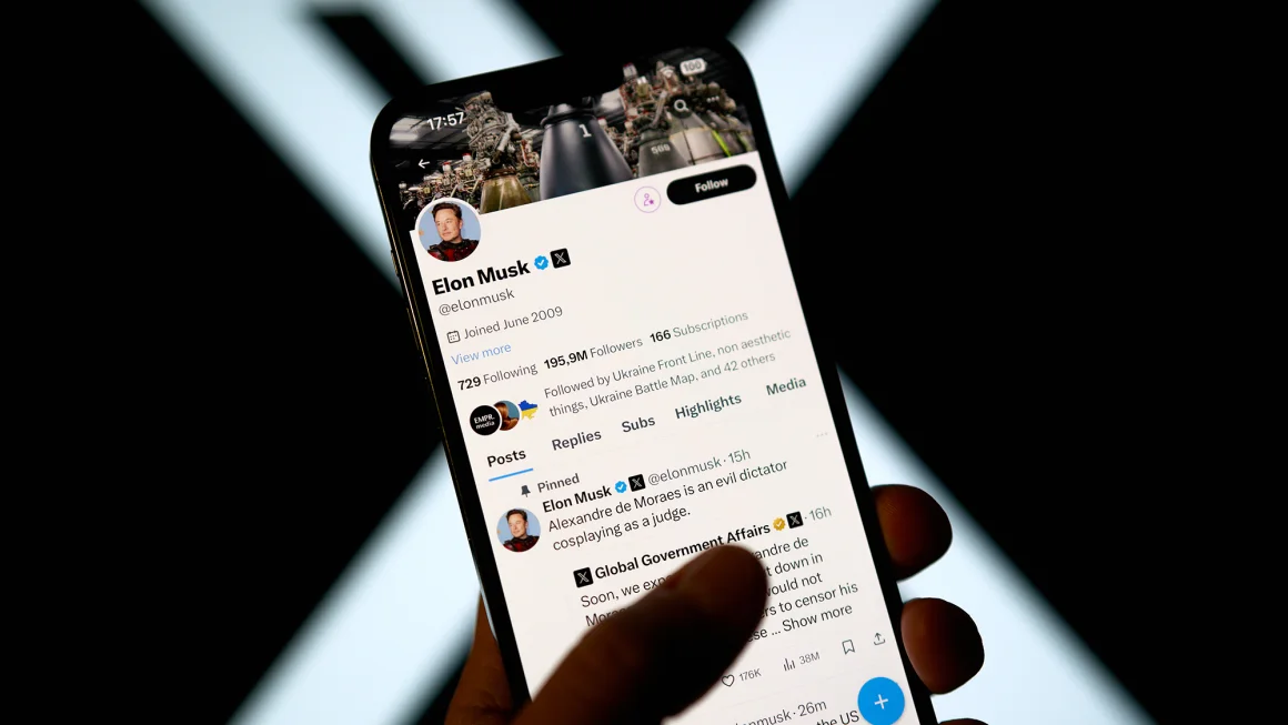 Captura de pantalla del perfil de Elon Musk en X (anteriormente Twitter) mostrada en un teléfono móvil. El perfil muestra la foto de Musk, su nombre de usuario @elonmusk, estadísticas de seguidores y publicaciones recientes. El fondo muestra el logotipo de X iluminado. Un dedo está tocando la pantalla del teléfono.