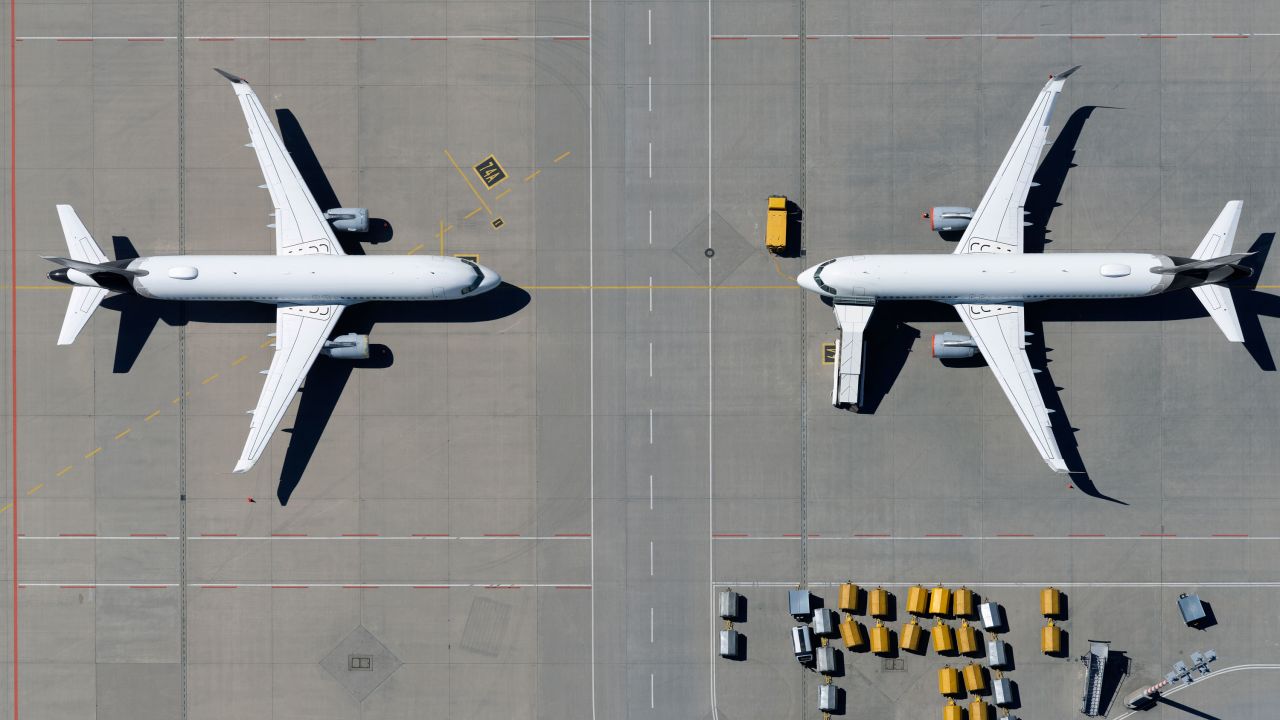 A view from above of airplanes parked face to face on sunny airport tarmac.