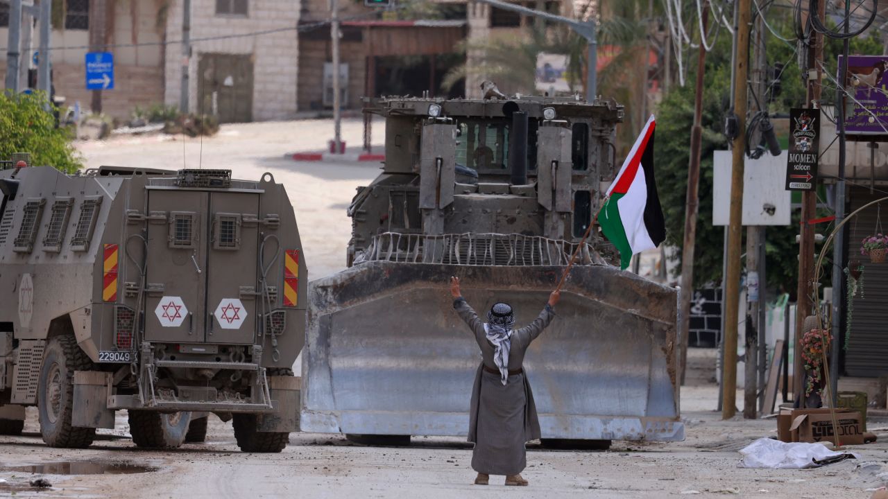 TOPSHOT - A Palestinian activist lifts a national flag and flashes the victory sign as Israeli armoured vehicles including a bulldozer drive on a street during a raid in Tulkarem on September 3, 2024, amid a large-scale military offensive launched a week earlier in the occupied West Bank. (Photo by Jaafar ASHTIYEH / AFP) (Photo by JAAFAR ASHTIYEH/AFP via Getty Images)