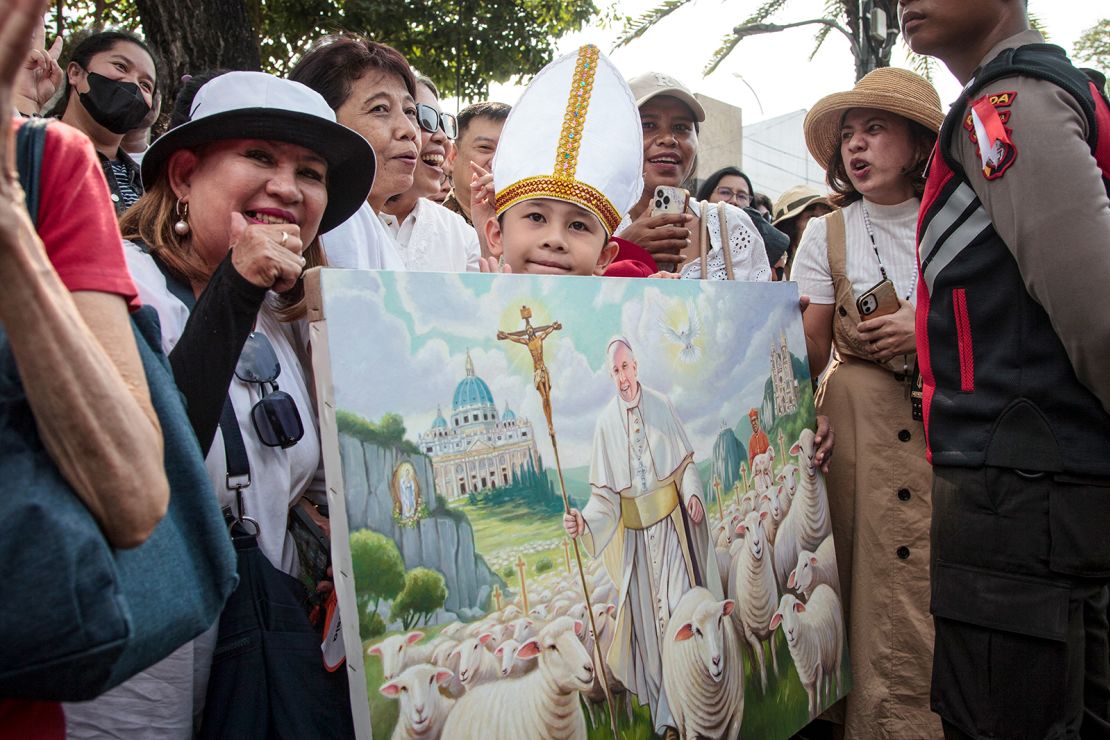 A boy holds a painting of Pope Francis as people wait before the appearance of Pope Francis on an electric screen in front of the Jakarta Cathedral in Jakarta on September 4, 2024.