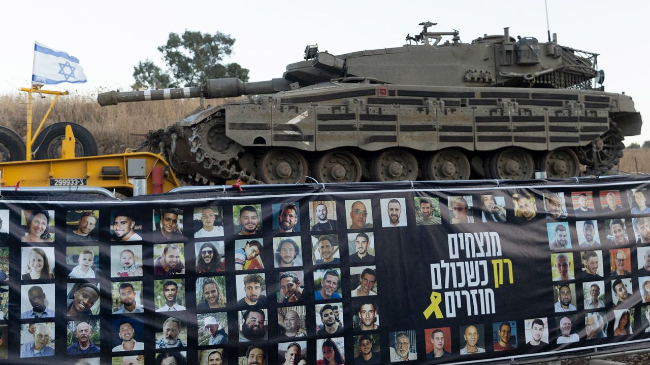 An Israeli tanks passes near a banner with photos of hostages held in the Gaza Strip, as it is transported on a truck towards the north of Israel on September 26, 2024.