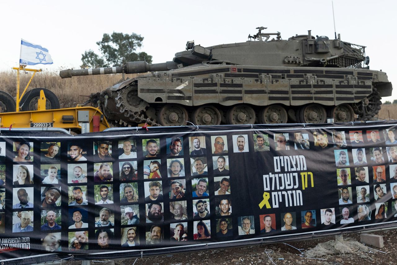 An Israeli tanks passes near a banner with photos of hostages held in the Gaza Strip, as it is transported on a truck towards the north of Israel on September 26, 2024.