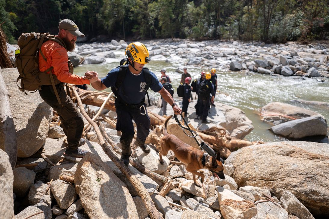 A member of FEMA urban search and rescue team New York Task Force One is assisted by a civilian search and rescue team member as they hike along the Broad River in the aftermath of Hurricane Helene near Chimney Rock, North Carolina, in October 2024.