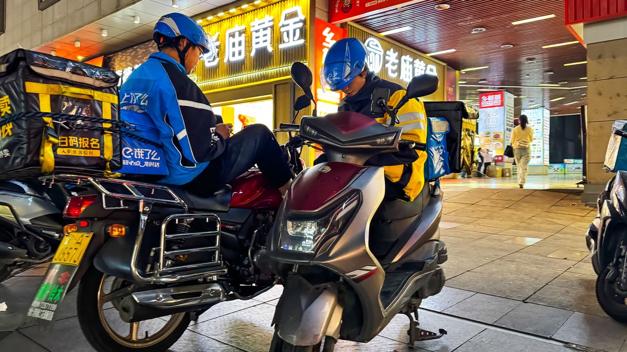 Two food delivery riders from different companies wait on their scooters for their next orders on October 7, 2024 in Chongqing, China.