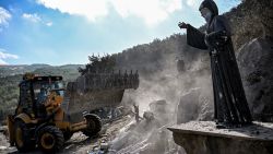 TOPSHOT - A Statue of the 19th-century Maronite Christian saint Mar Charbel is pictured as a bulldozer moves to clear rubble and debris from the site of a previous Israeli air strike on the village of Aito in northern Lebanon on October 15, 2024. The Lebanese Red Cross said at least 18 people died in a strike on north Lebanon on October 14, with the health ministry and official media reporting an Israeli raid on the Christian-majority area far from Hezbollah strongholds. (Photo by JOSEPH EID / AFP) (Photo by JOSEPH EID/AFP via Getty Images)