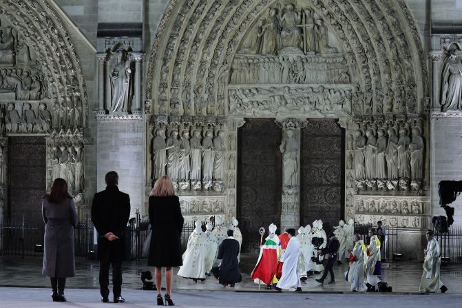 Shortly after sunset, Paris Mayor Anne Hidalgo, left, French President Emmanuel Macron and his wife Brigitte Macron stand in the refurbished forecourt, as the Archbishop of Paris Laurent Ulrich leads a procession of archbishops, bishops and priests to the cathedral doors at the start of the ceremony.