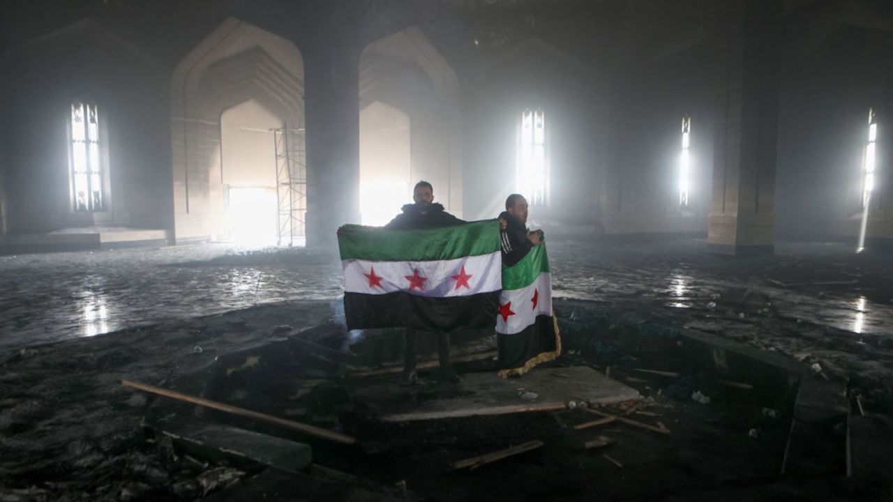 TOPSHOT - Rebel fighters stand with the flag of the revolution on the burnt gravesite of Syria's late president Hafez al-Assad at his mausoleum in the family's ancestral village of Qardaha in the western Latakia province on December 11, 2024, after it was stormed by opposition factions and set ablaze. The father and predecessor of Syria's ousted president Bashar al-Assad died in 2000. (Photo by AAREF WATAD / AFP) (Photo by AAREF WATAD/AFP via Getty Images)