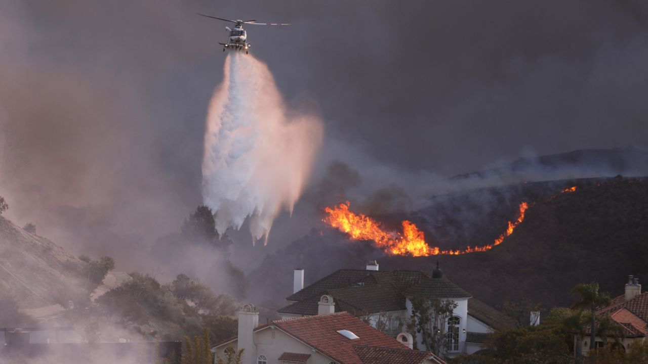 A helicopter drops water around homes threatened by the wind-driven Palisades Fire in Pacific Palisades, California, January 7, 2025. A fast-moving brushfire in a Los Angeles suburb burned buildings and sparked evacuations Tuesday as "life threatening" winds whipped the region. More than 200 acres (80 hectares) was burning in Pacific Palisades, a upscale spot with multi-million dollar homes in the Santa Monica Mountains, shuttering a key highway and blanketing the area with thick smoke. (Photo by David Swanson / AFP) (Photo by DAVID SWANSON/AFP via Getty Images)